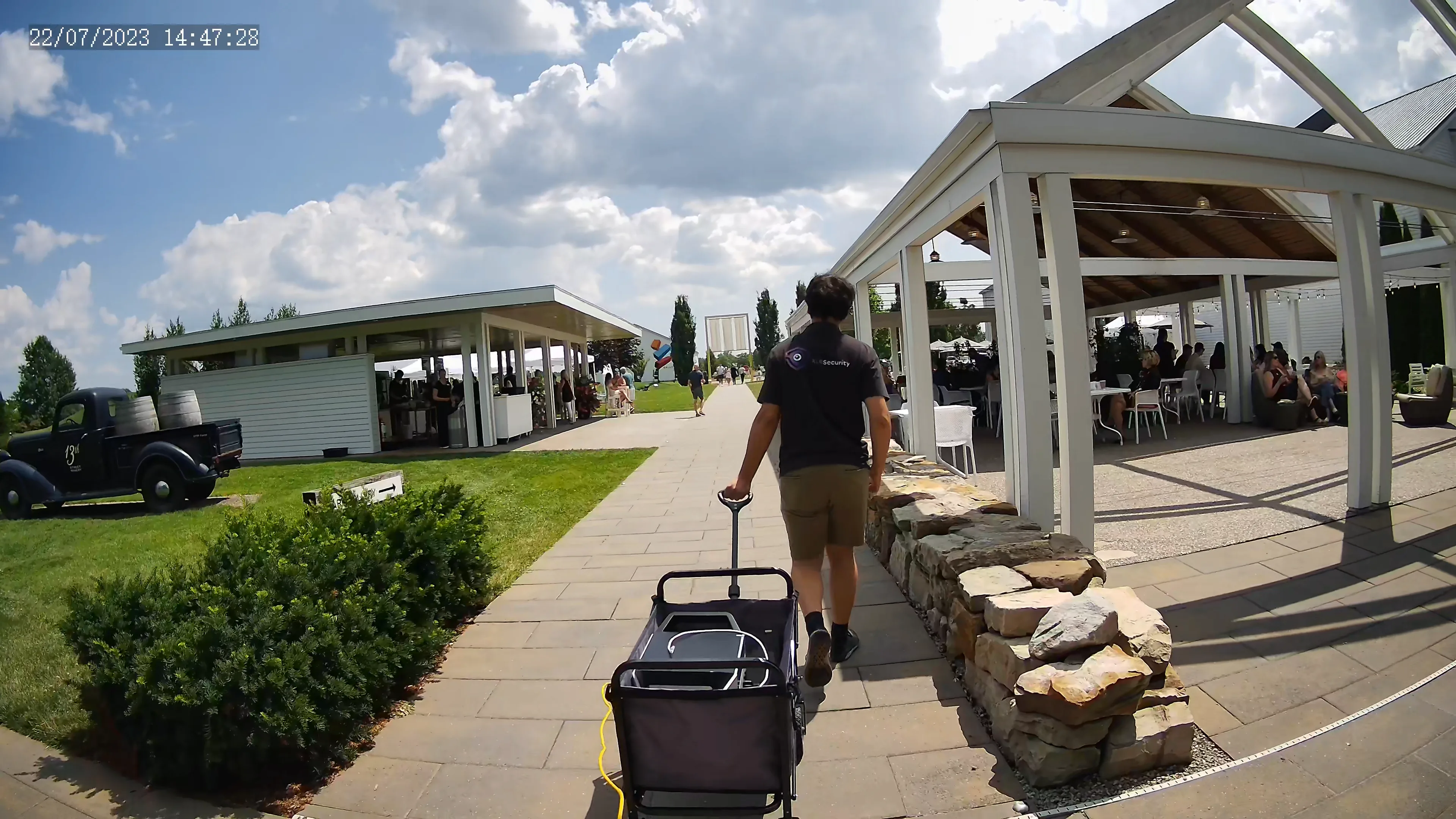 A young man pulling a cart, walking towards a winery with many people enjoying lunch outside