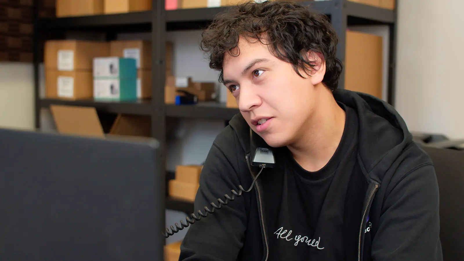 A man on a phone call with the phone held between his shoulder and cheek. He is staring at a computer monitor, most likely doing tech support.