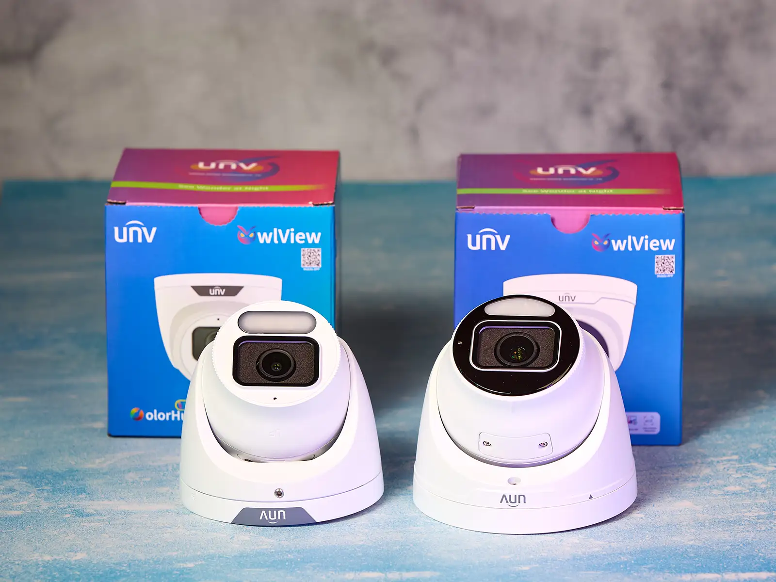 Uniview's 8MP OwlView and OwlView Plus cameras on a table with a blue background. Both of the camera boxes are behind the cameras.