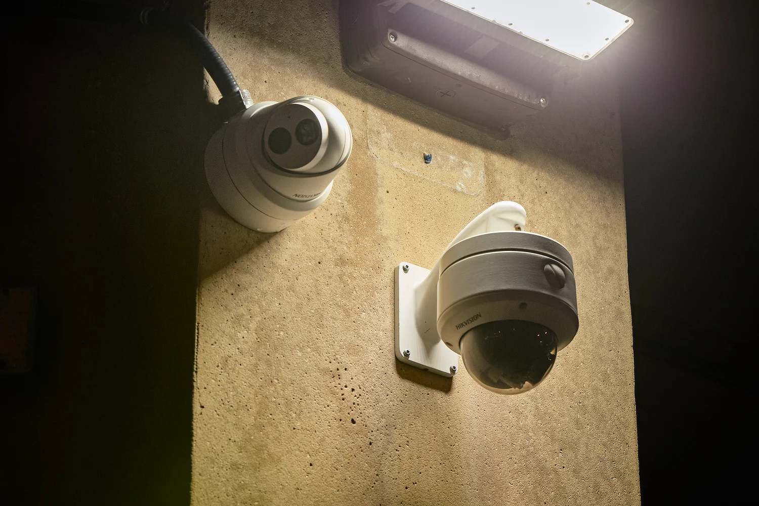 A Hikvision turret and dome camera mounted on a concrete wall beneath a streetlight at night