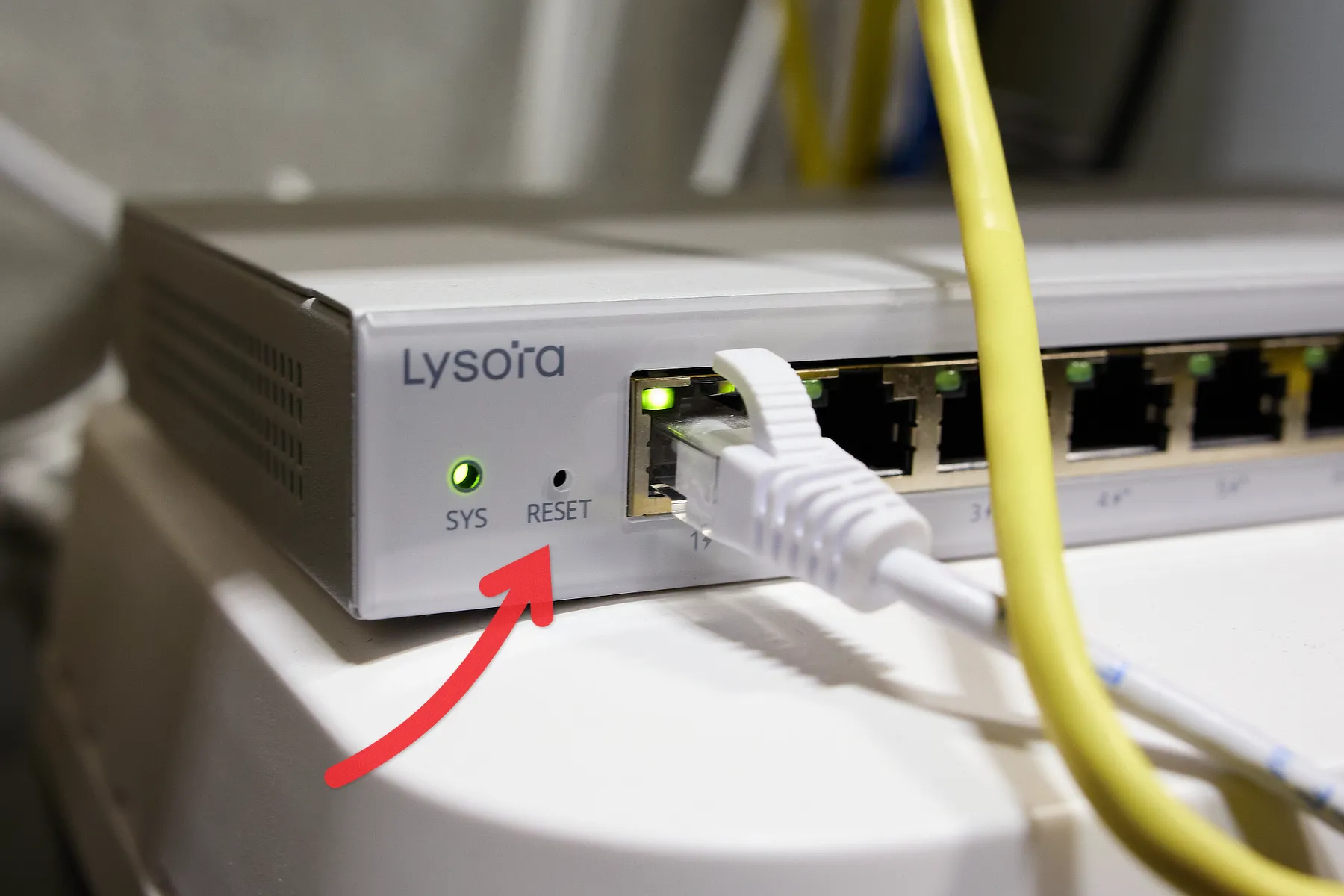 A light-grey router with the text Lysora printed on it, along with a red arrow pointing to the reset button.