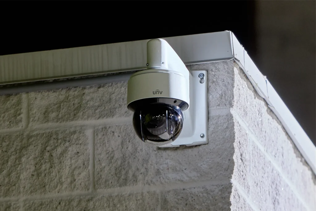 A Uniview dome PTZ camera, mounted on a wall mount bracket to a white brick wall against the night sky in the background.
