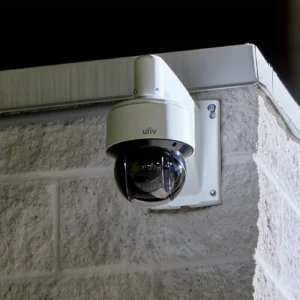 A Uniview dome PTZ camera, mounted on a wall mount bracket to a white brick wall against the night sky in the background.