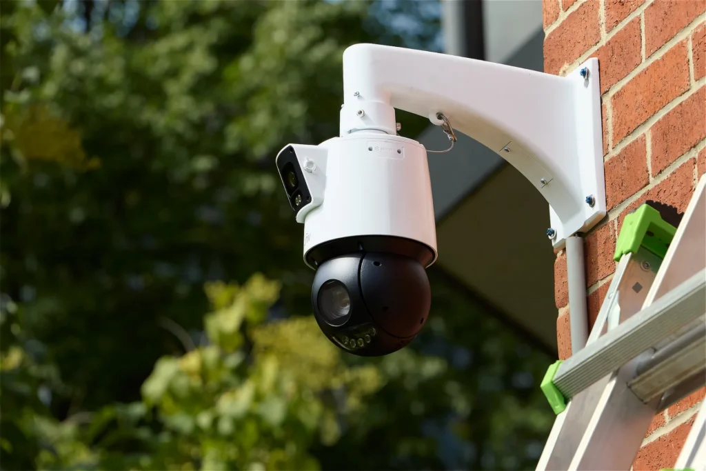 A large Uniview PTZ camera mounted on a red brick wall, with trees in the background and a ladder resting against the wall at the bottom right corner of the photo.